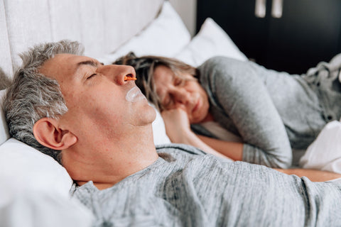 A couple sleeps in bed. The man is wearing a nasal dilator and SomniFix mouth tape to prevent snoring and boost sleep quality.