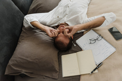 A person lays on a bed surrounded by notebooks, rubbing their eyes due to being tired.