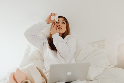 A woman sits in bed in front of her laptop and applies eye drops to her eyes.