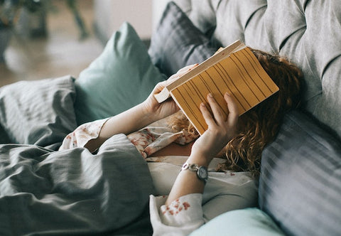 A woman lays in bed sleeping with a book covering her face.