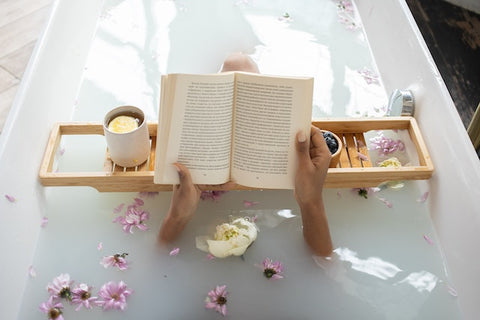 A woman sits in a hot bath filled with flowers with tea nearby while reading a book.