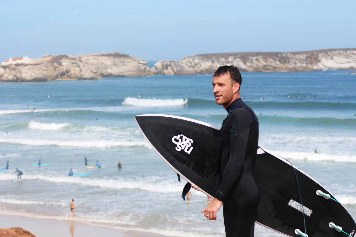 ben holding a black surfboard with a white cskins sticker is wearing a black wetsuit looking at the view