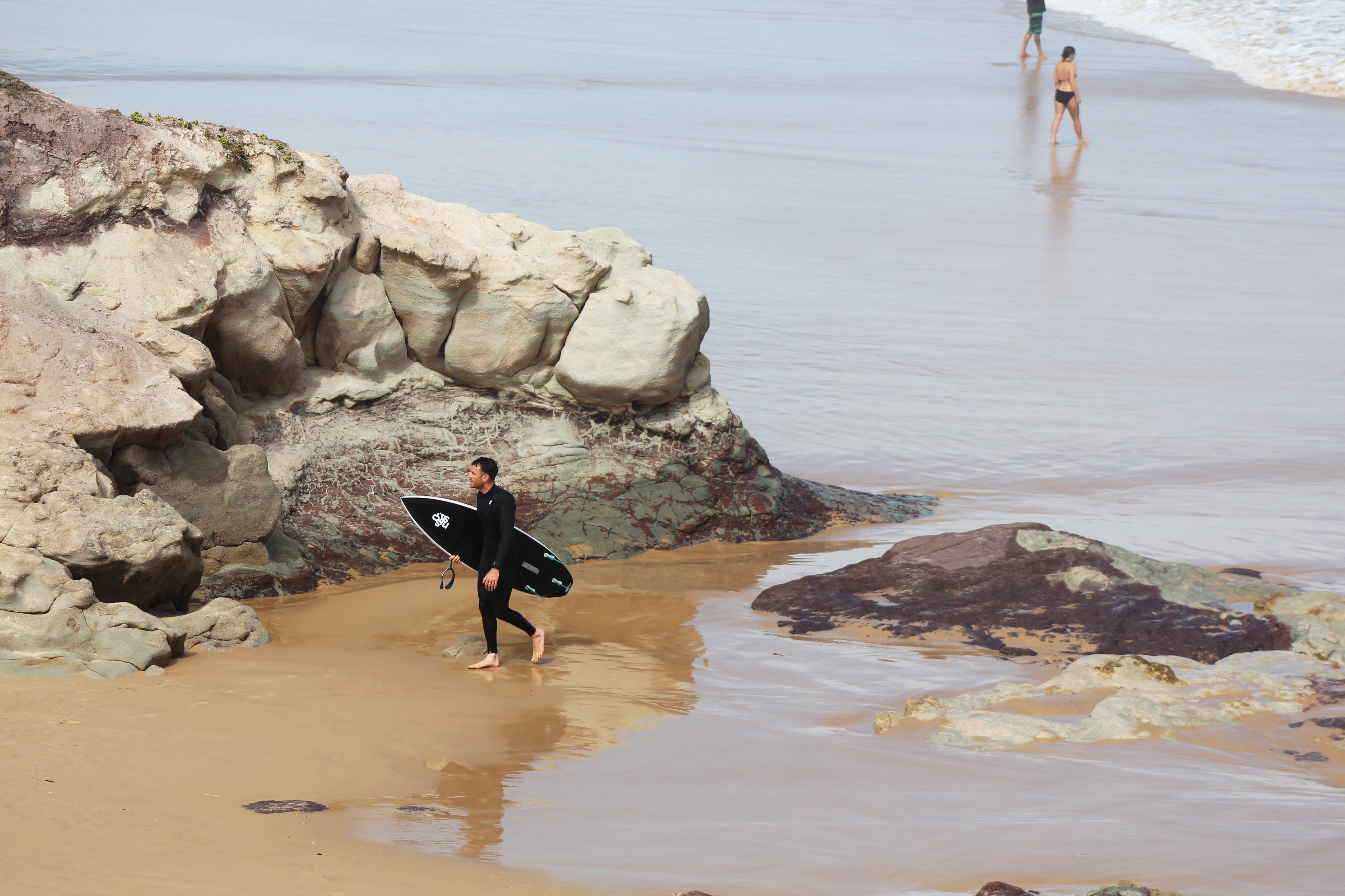 ben exits the water wearing a skins wetsuit holding a black surfboard