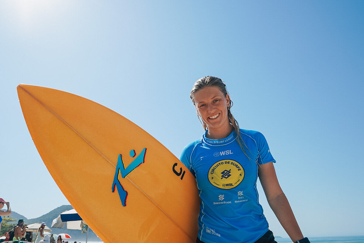 Sophie smiling wearing a blue rash vest holding an orange surfboard