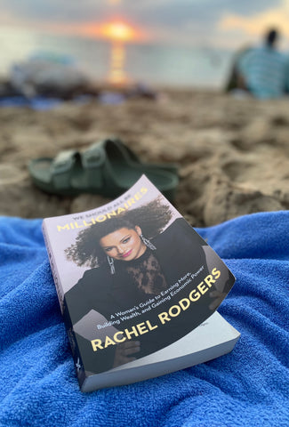 Picture of Book We should all be millionairs by Rachel Rodgers. The book is on a blue towel with the sunset and beach behind it.