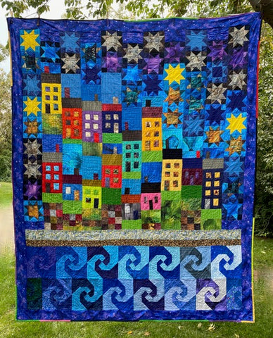 Jellybean Quilt with colourful houses set between a sky of quilted block featuring stars and and waves based on the Jellybean Row of downtown St. John's is shown against a garden background.