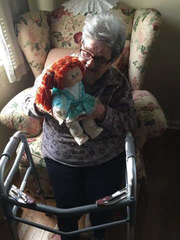 An old woman sits in a chair with a walker, holding a red haired cabbage patch doll
