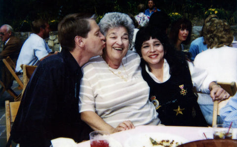 A grandmother sits betweeen her son in law and daughter. Son in law is giving her a kiss on the cheak