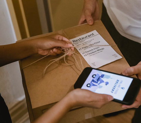 Black android phone and food in brown boxes being exchanged in a doorway between a customer and a delivery person.