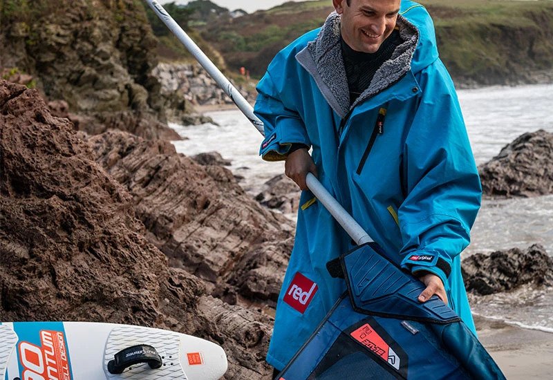 Man wearing blue changing robe pulling paddle board paddle from a paddle bag