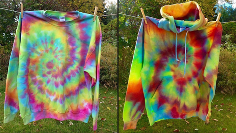 Two different tie dye hoodies side-by-side hanging on a clothesline outdoors. To the left is a neon rainbow spiral crewneck sweatshirt, on the right is a red, yellow & blue spiral tie-dyed pullover hoodie