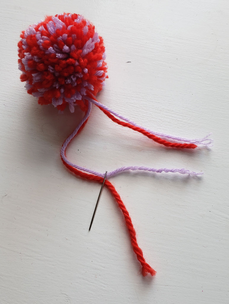 A red and purple pom pom with thread tails and a hand sewing needle