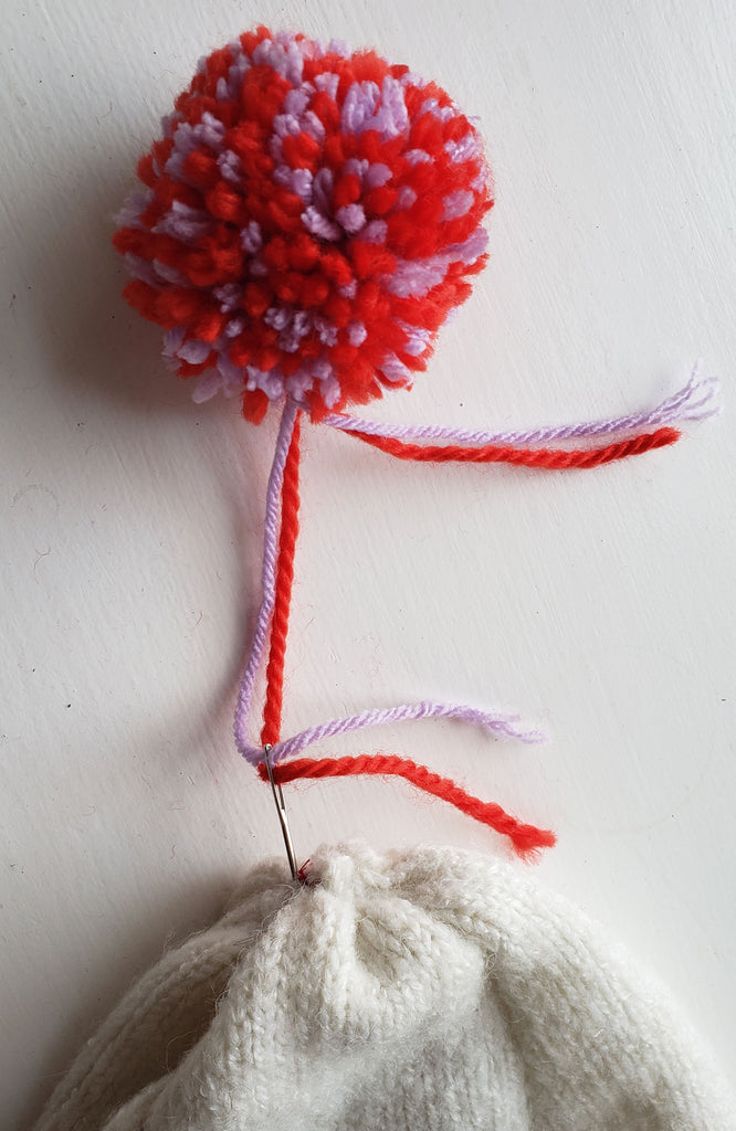 Reb and purple pom pom being attached by a hand sewing needle to the top of a winter hat
