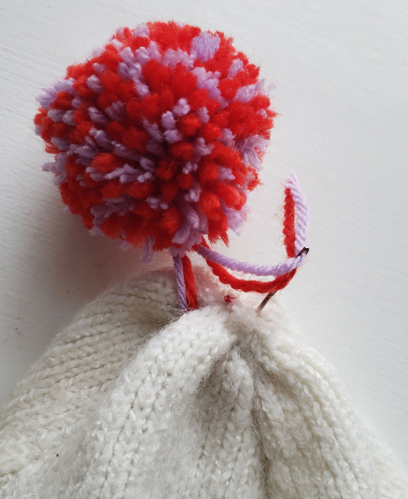 Yarn being sewn through to the wrong side of a winter hat, securing a pom pom