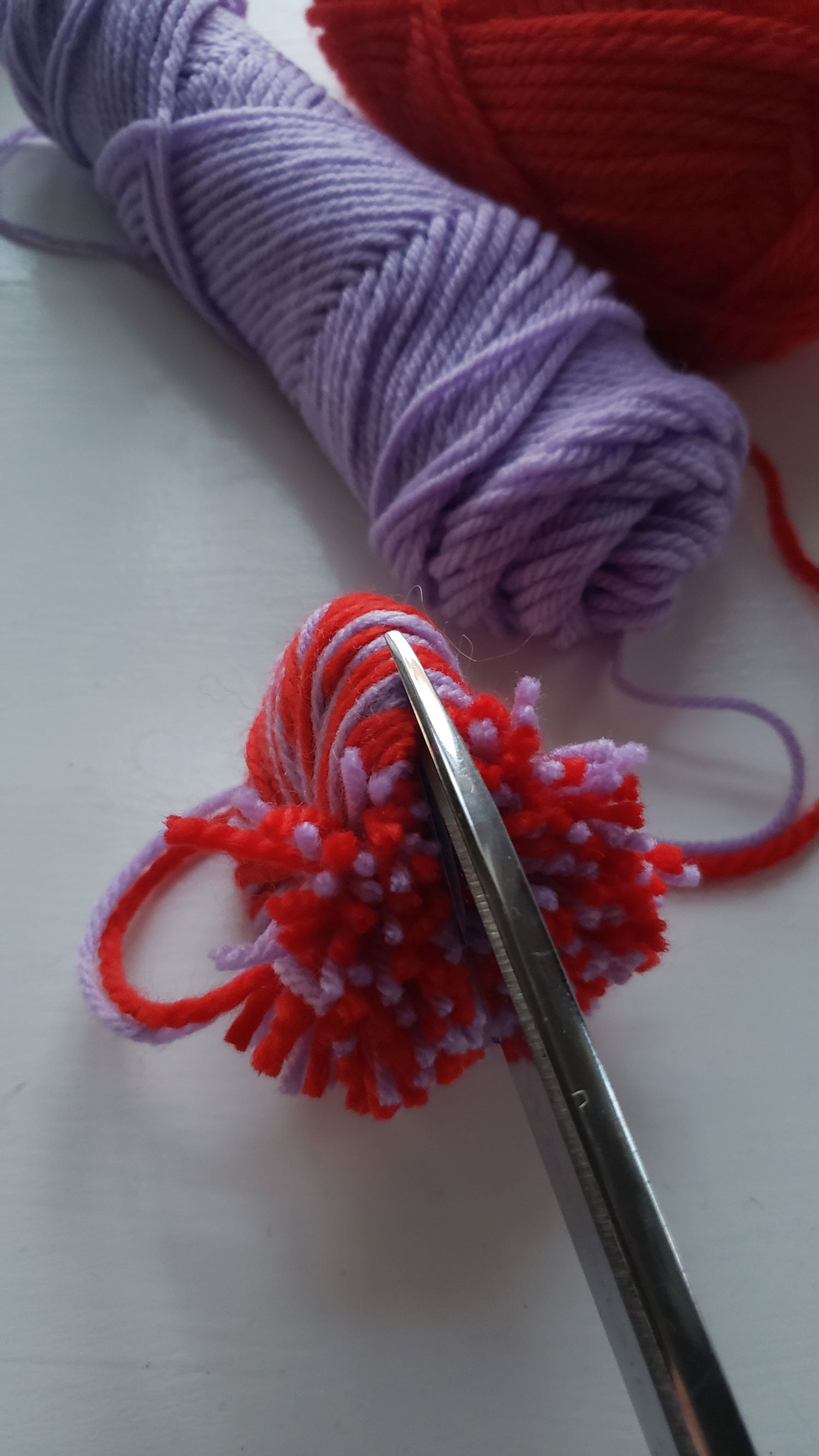 A scissors cuts yarn along the edge of a pom pom maker