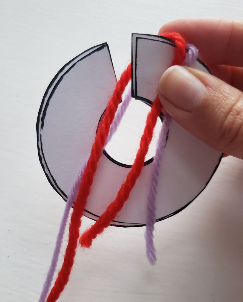 Red and purple yarn is being wrapped around a pom pom disk