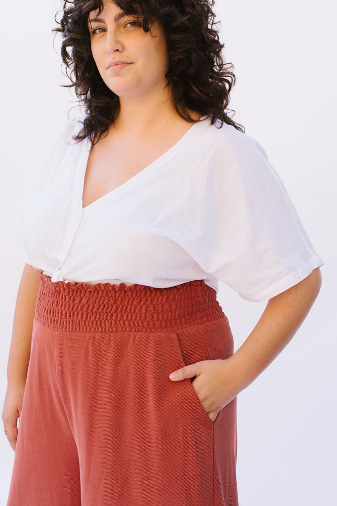 A woman wearing a white top and coral coloured pants with a shirred waistband. Her hand is in the side seam pocket
