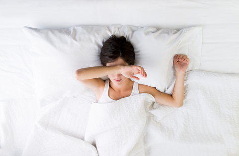 woman in bed covering her eyes with part of her arm laying down on white linen bedding