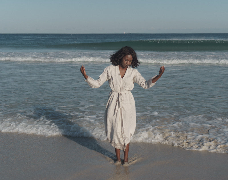 Black woman standing in front of the ocean on the sand. She is looking down and away from the camera.