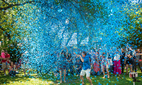 Couple posing covered with blue confetti to announce having a baby boy while surrounded by friends and family.