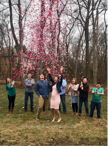 Couple posing, surrounded by family and friends outside while shoot pink confetti in the air