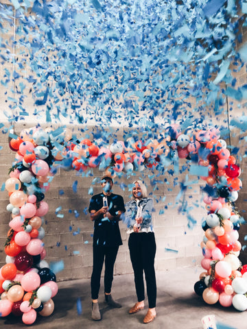 Man and Woman surrounded by colorful balloons while shooting blue confetti cannons.