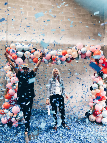 Couple celebrating after shooting blue confetti cannons. While surrounded by colorful balloons.
