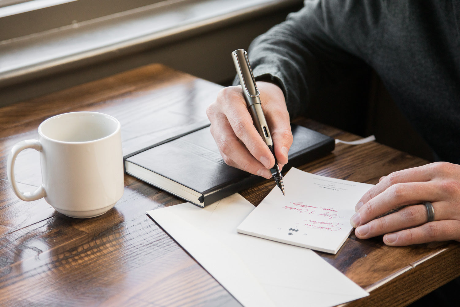 hand holding a fountain pen at a wooden desk with notebook and paper