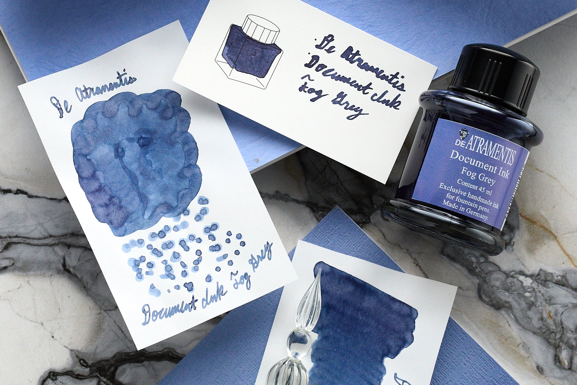 45 mL glass bottle of De Atramentis Document Ink Fog Grey Fountain Pen Ink and 3 white cards with writing and doodles using Document Ink Fog Grey on marble background with blue notebooks.