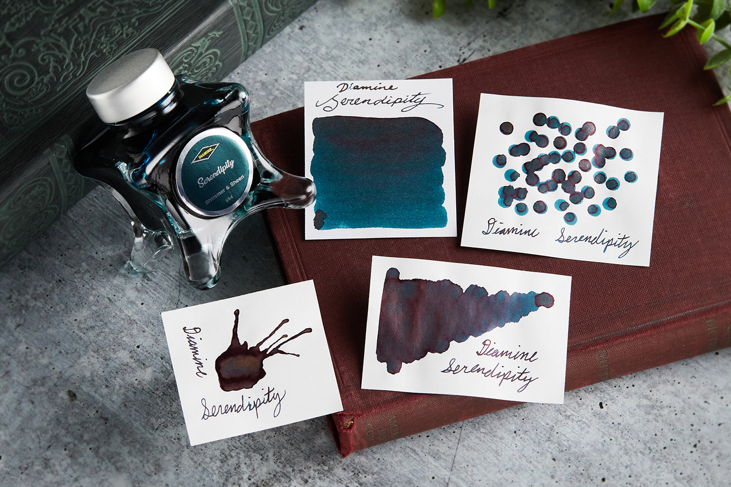 Diamine Serendipity fountain pen ink bottle along with 4 white cards that have ink swabs, splatters, or dots, all resting on a brown notebook with a gray background