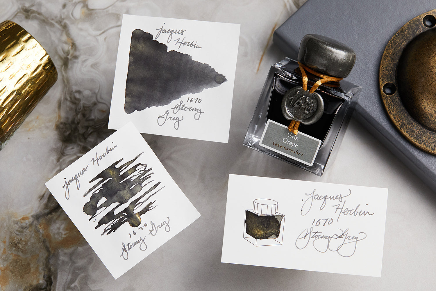 Jacques Herbin 1670 Stormy Grey fountain pen ink bottle with three white cards that have scatchy blob, tornado, and blob on them, all on light colored background