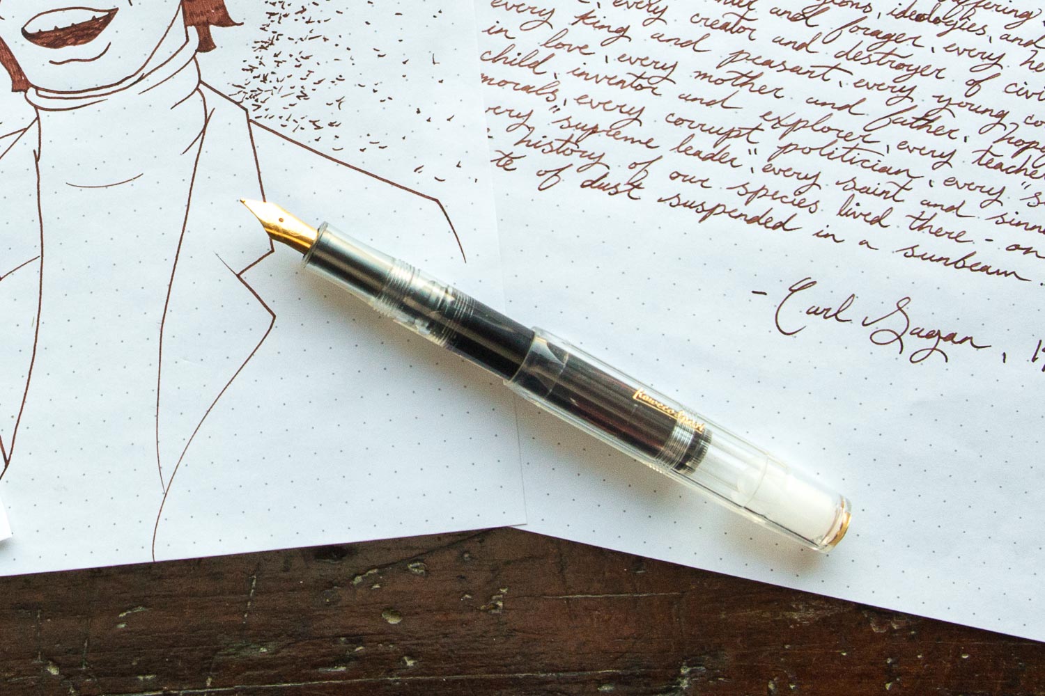 A clear fountain pen filled with ink, on top of a drawing and writing