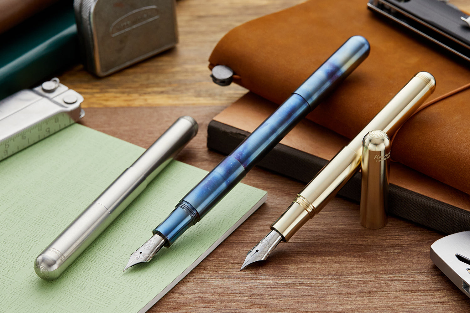 Kaweco Supra fountain pens in brass, steel and flame blue. Capped and uncapped on wooden desk