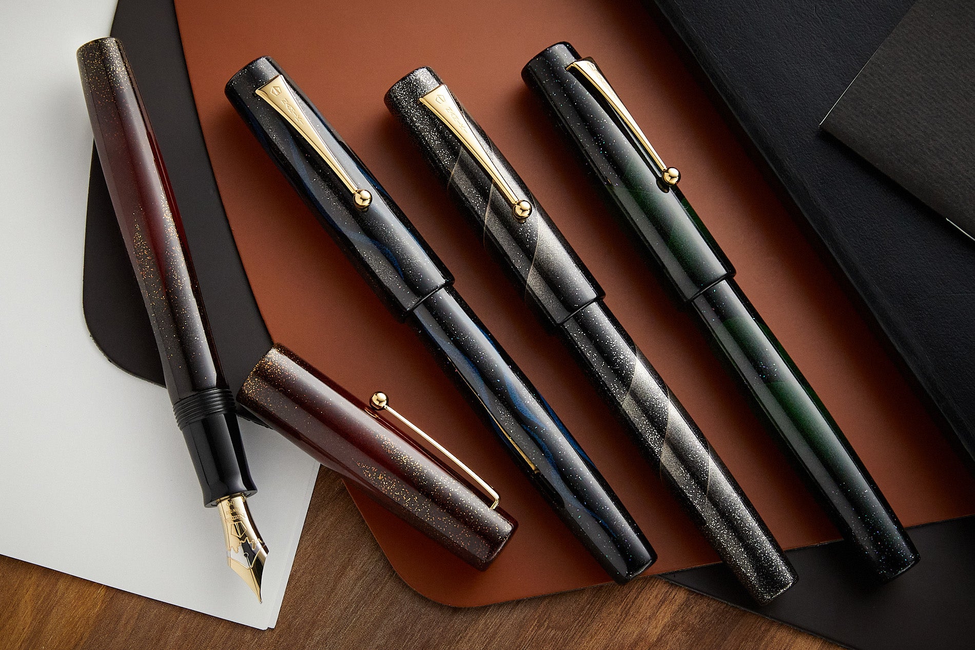 Four Namiki Aya Maki-e fountan pens on a leather writing matt and desk background. Pens are in assorted designs, all muted colors