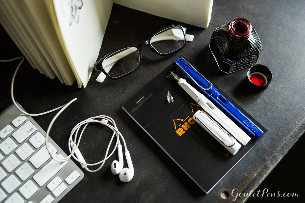 Several fountain pens on top of a small notebook, with glasses and an open bottle of ink, on a desk