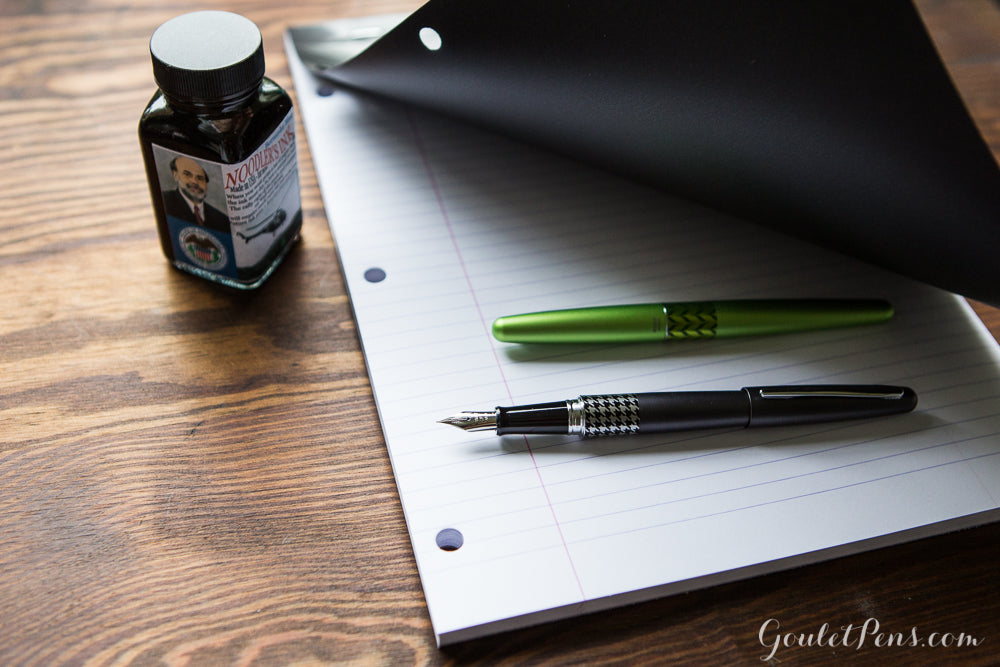 Several fountain pens peeking out under the cover of a lined notepad