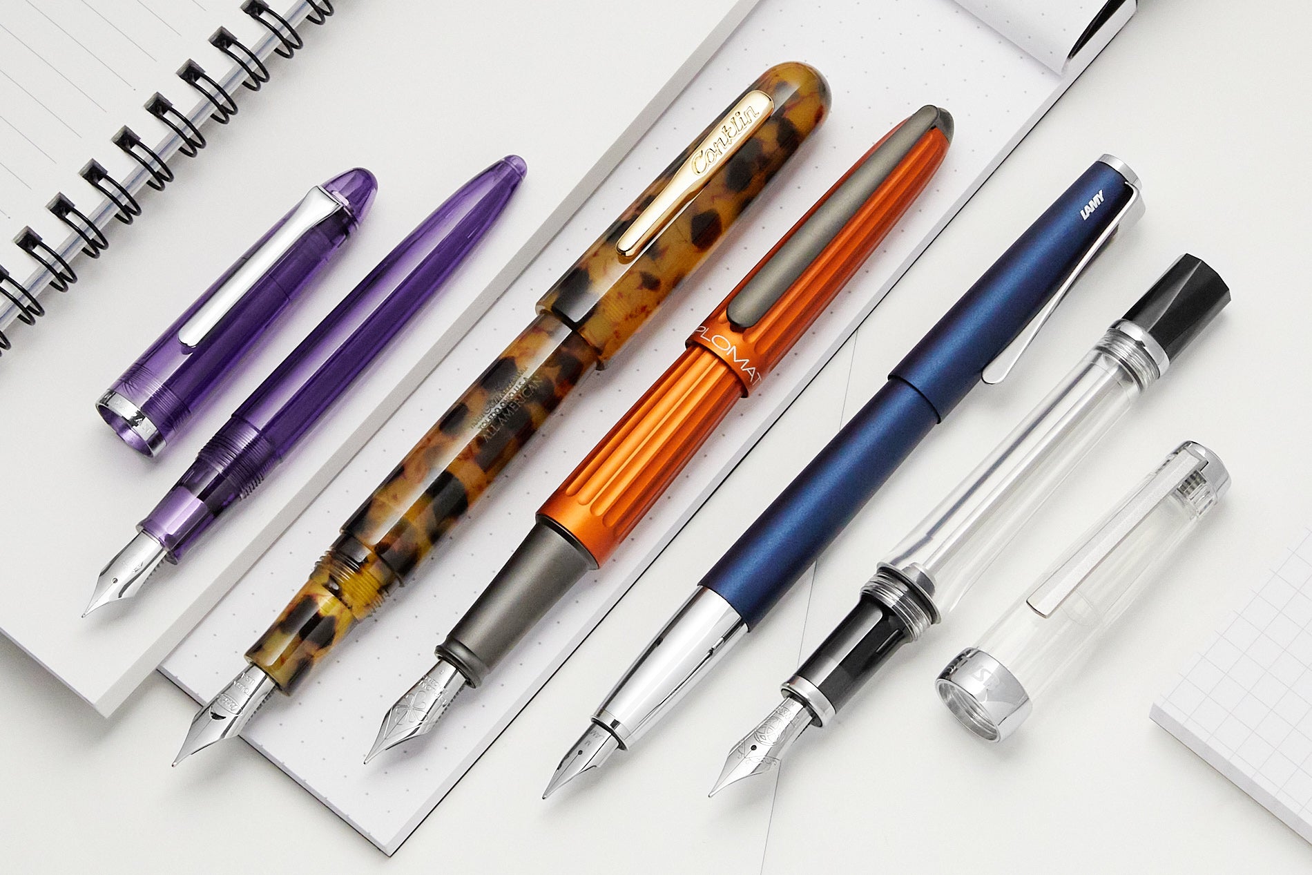 A group of open fountain pens lined up diagonally on top of open notebooks