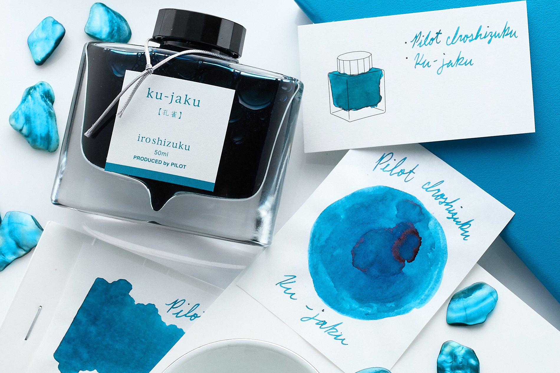 Pilot Iroshizuku Ku-Jaku Fountain Pen Ink writing sample and swaps with bottle on desk type background and bright blue bits of abalone shell as accents