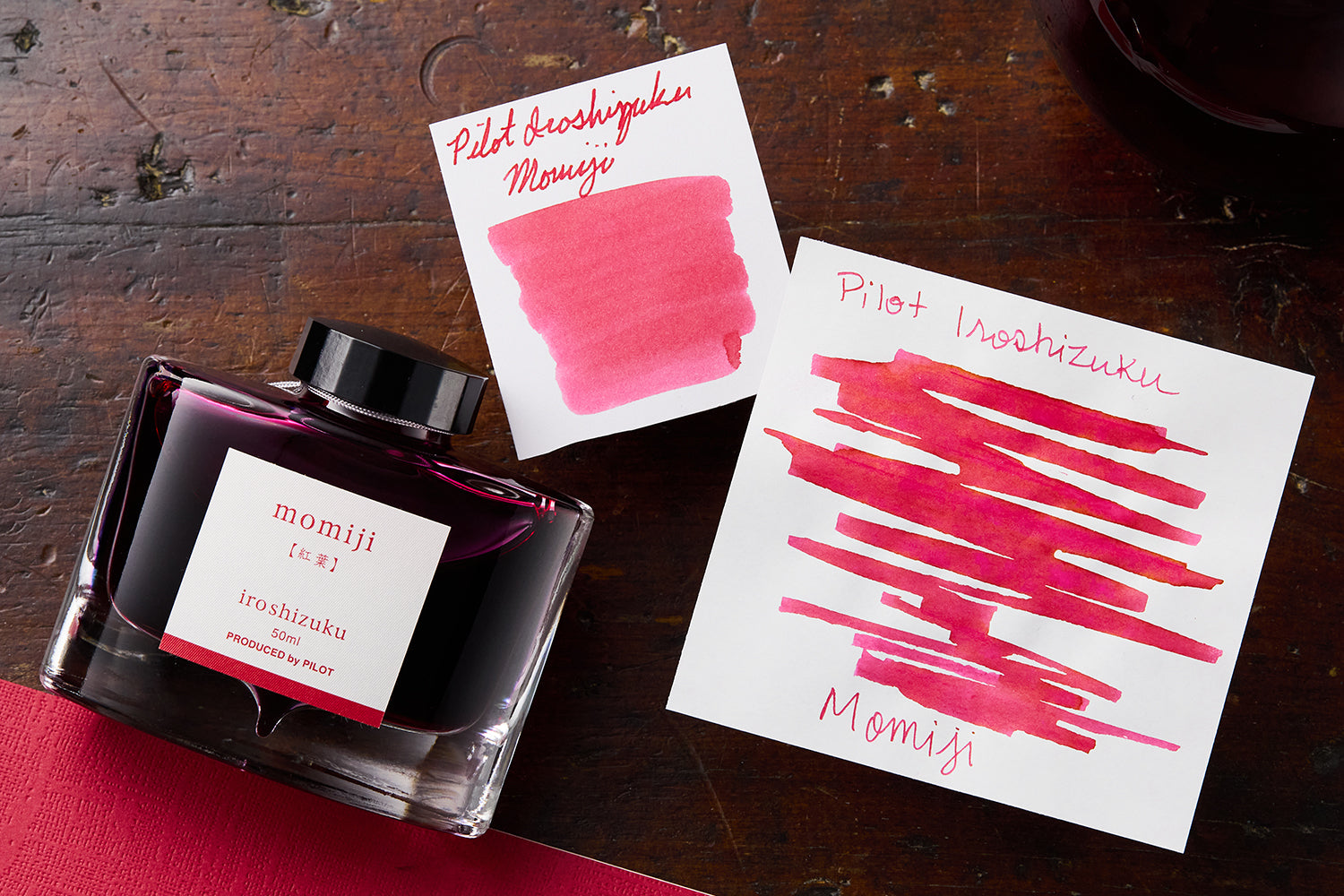 Pilot Iroshizuku Momiji fountain pen ink bottle, and 2 white cards with pink ink swabs on them on wooden background