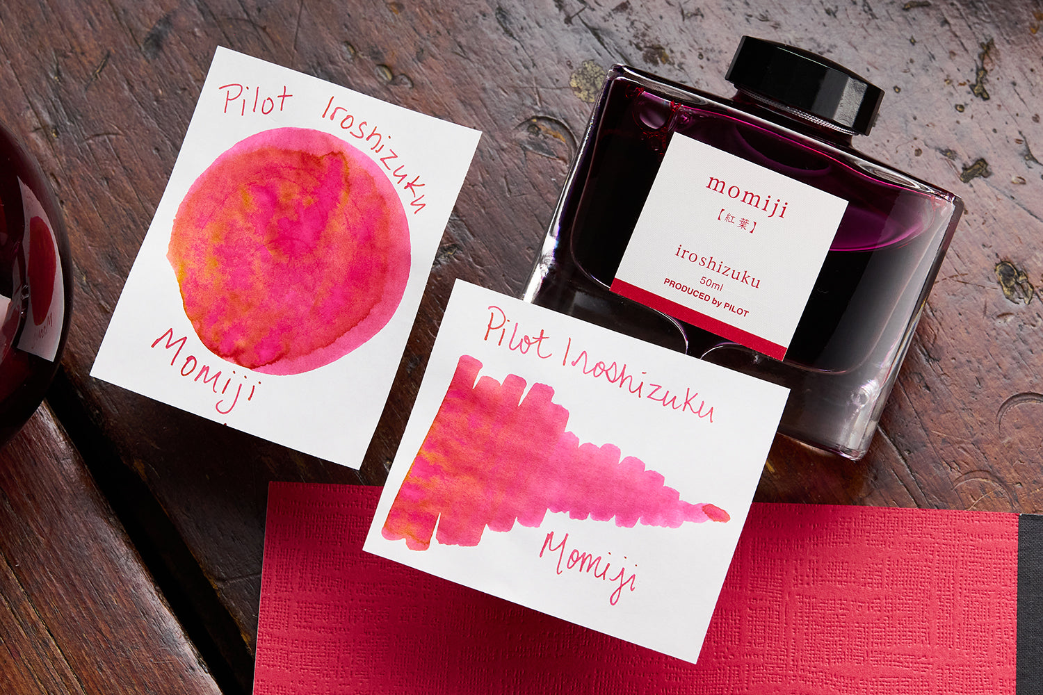 Pilot Iroshizuku Momiji fountain pen ink bottle, and 2 white cards with pink ink swabs on them on wooden background