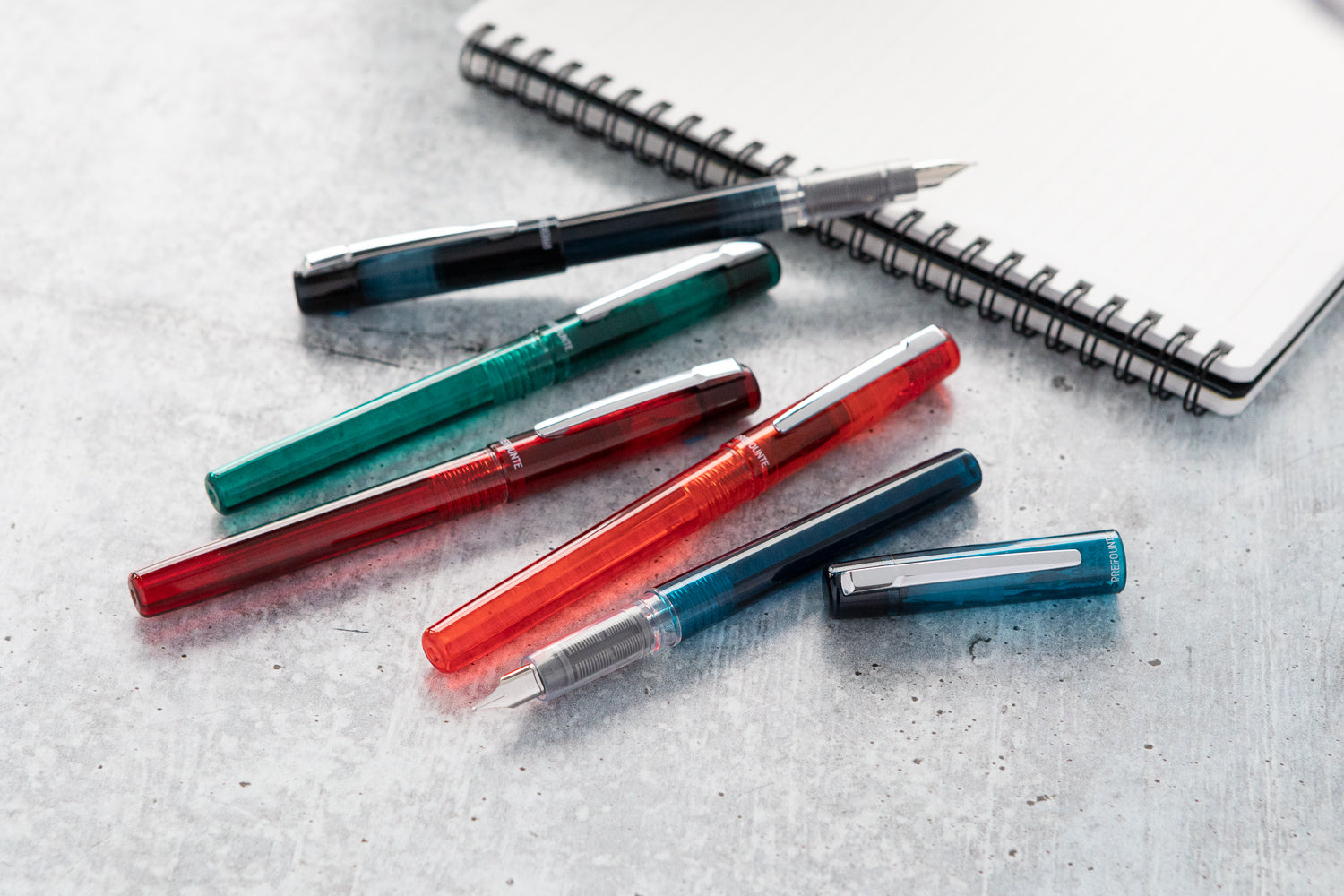 A group of five fountain pens scattered on a grey marble background with a notebook