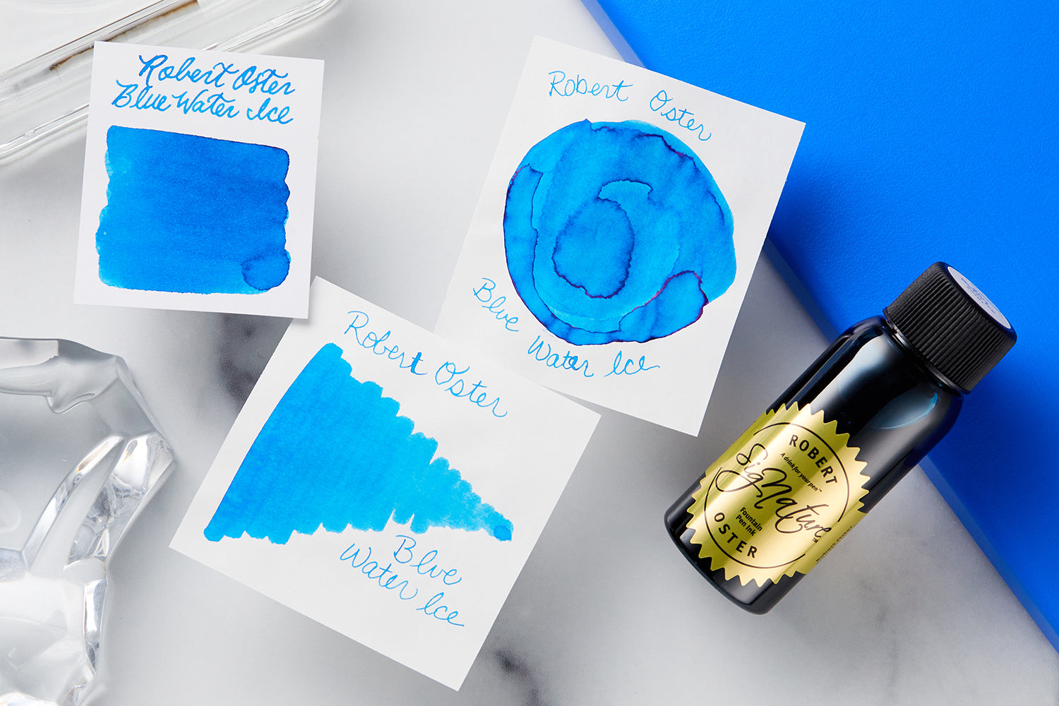 Robert Oster Blue Water Ice Fountain Pen ink bottle, 2 swatches and a swab on marbled desk background