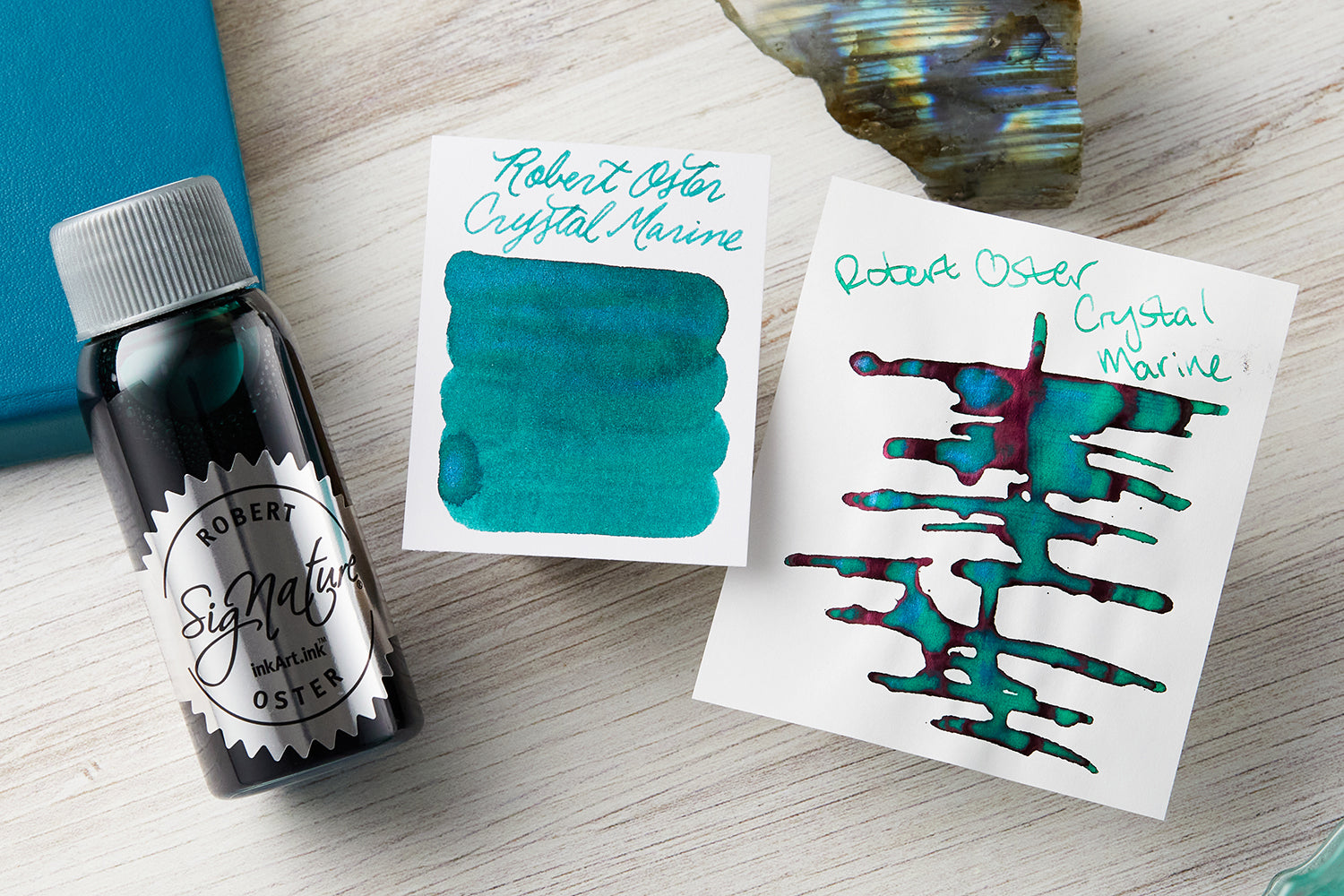 Robert Oster Fountain Pen ink bottle with 2 ink swab cards with a teal journal in the background