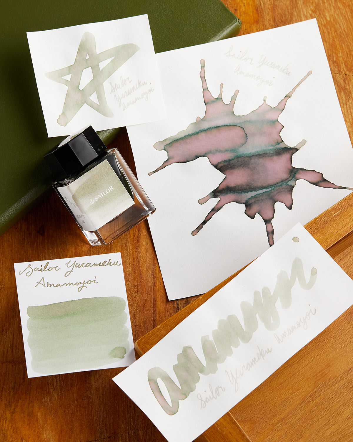 Four white pieces of paper with Sailor Yurameku Amamoyoi Fountain Pen ink on each card in a different shape or pattern and glass product bottle in between them.