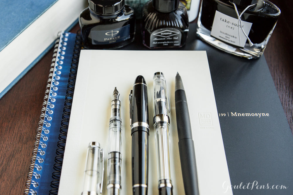 Assorted fountain pens uncapped on a notebook with ink bottles around the desk