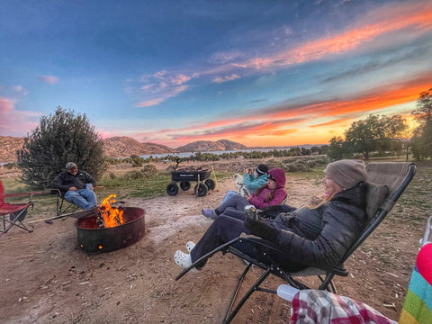 campers sit around a fire with sunset and mountains in the background