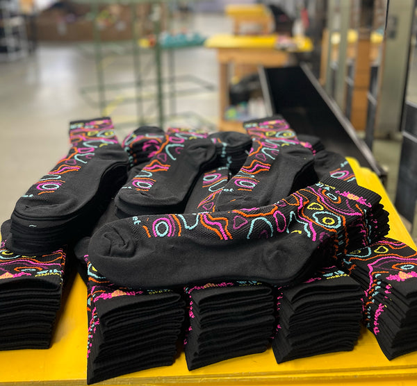 black custom knit socks with a colorful topographic design are stacked on a board in the factory