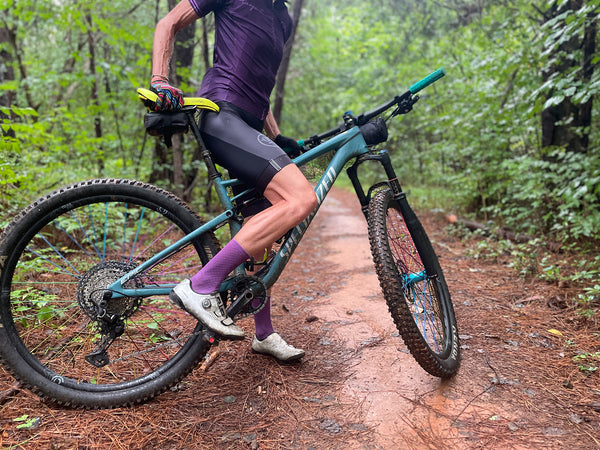 female cyclist on a mountain bike on a wooded trail wearing purple socks to match her kit