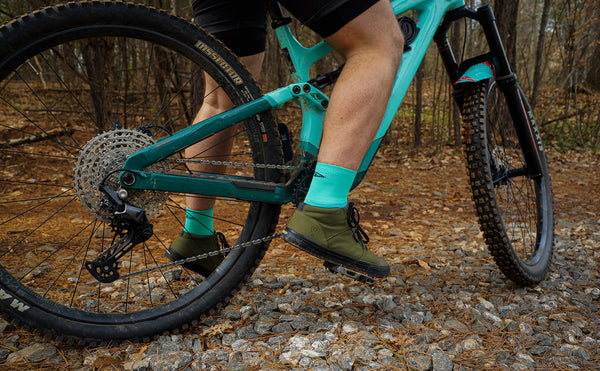 a male cyclist sits on a bike in the woods wearing teal DeFeet cycling socks that match his bike