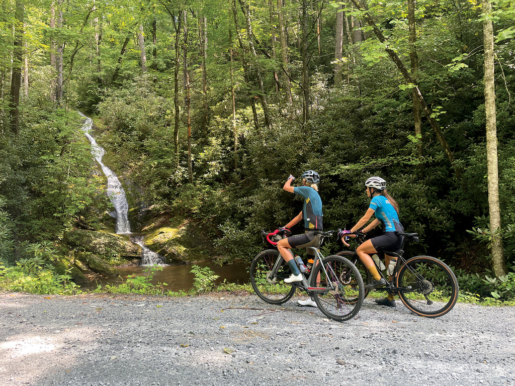 two female cyclists on bikes on a gravel road facing a waterfall, both wearing socks that match their kits